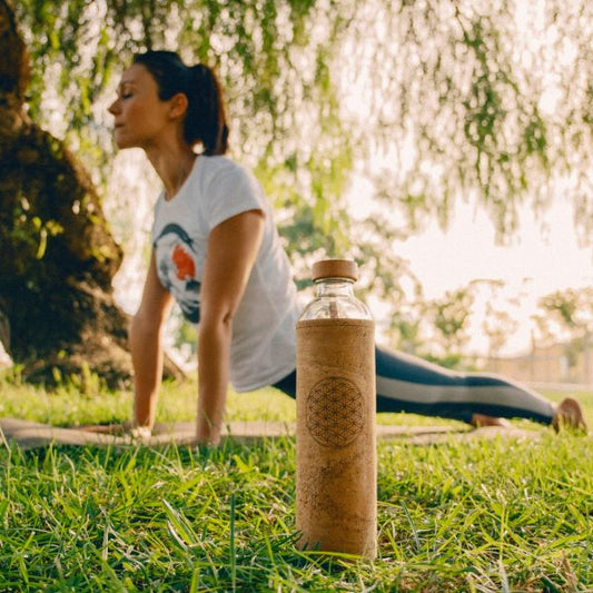 Mujer practica yoga para su bienestar con una botella de vidrio con funda de corcho y diseño de geometría sagrada.