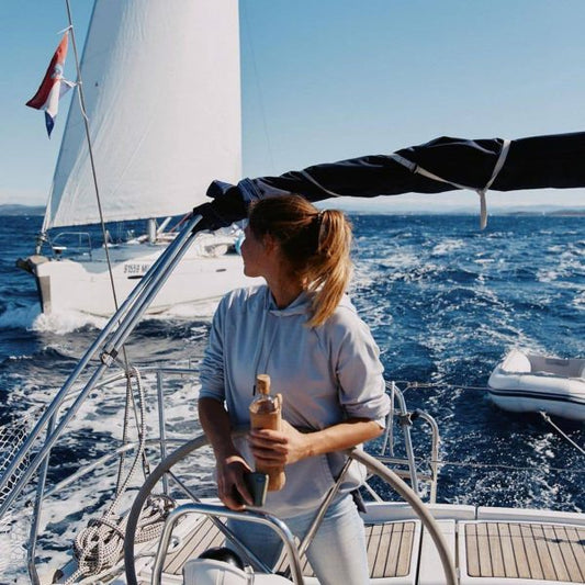 Persona al timón de un velero navegando, con una botella de vidrio en la mano para su hidratación.