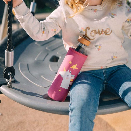 Niña en un columpio de un parque junto a su botella de vidrio con una funda rosa de unicornio para su hidratación.