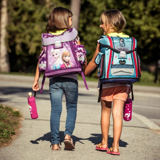 Dos niñas con mochilas caminan de la mano, llevando sus botellas para una correcta hidratación.