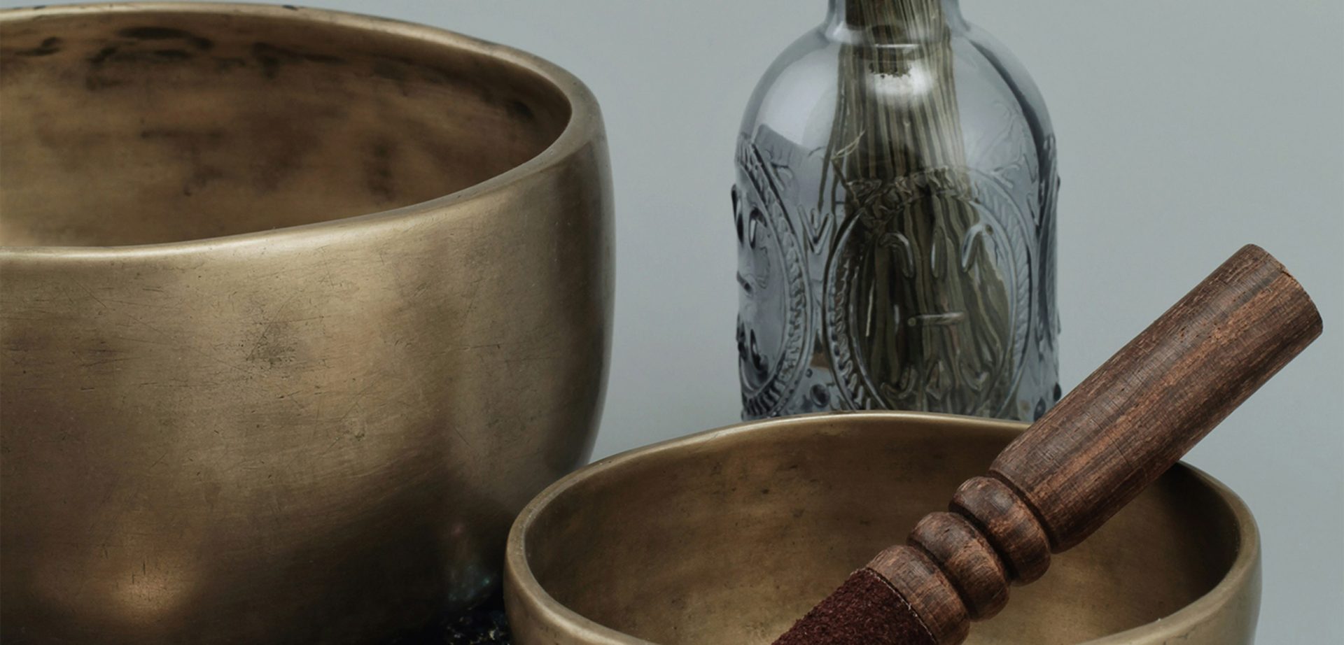 Dos cuencos tibetanos de bronce, un mortero de madera y una botella de vidrio ornamentada sobre un fondo liso.