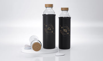 Sobre una plataforma blanca se exhiben dos botellas de cristal Flaska con tapón de madera y funda de neopreno negras con diseño de signos zodiacos Libra y Escorpio.