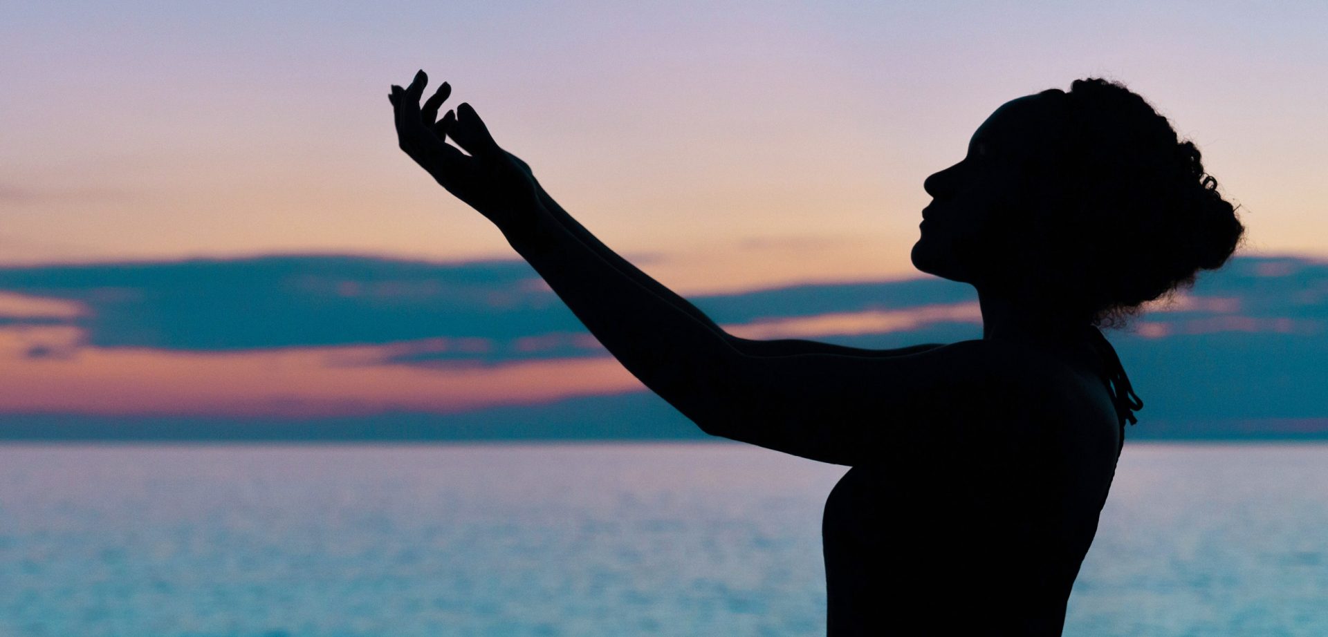 Silueta de una persona con las manos en alto frente a una tranquila puesta de sol junto al mar, con un cielo colorido y un océano en calma de fondo. En primer plano, un una botella de cristal Flaska, lo que contribuye a la serenidad del entorno.