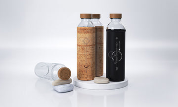 Tres botellas de vidrio con funda de corcho y funda de neopreno negra sobre una bandeja redonda, rodeadas de pequeñas piedras sobre fondo blanco.