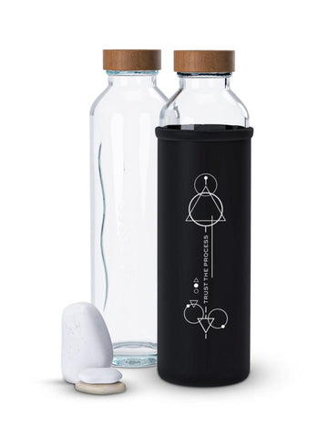 Dos botellas de vidrio con tapa de madera, protegida con una funda negra de neopreno con diseños geométricos y texto, colocada junto a tres piedras.